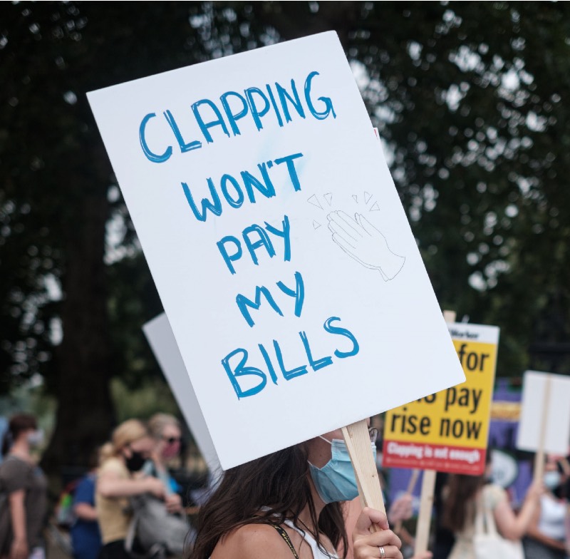 Main image: Nurse at protest holding sign reading 'Clapping won't help pay my bills' (Ehimetalor Akhere Unuabona/Unsplash)